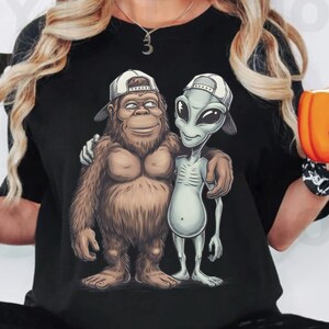 May include: Black t-shirt featuring a cartoon illustration of a Bigfoot character and an alien, both wearing white baseball caps. The Bigfoot is brown and furry, while the alien is gray with large eyes. They are embracing each other.