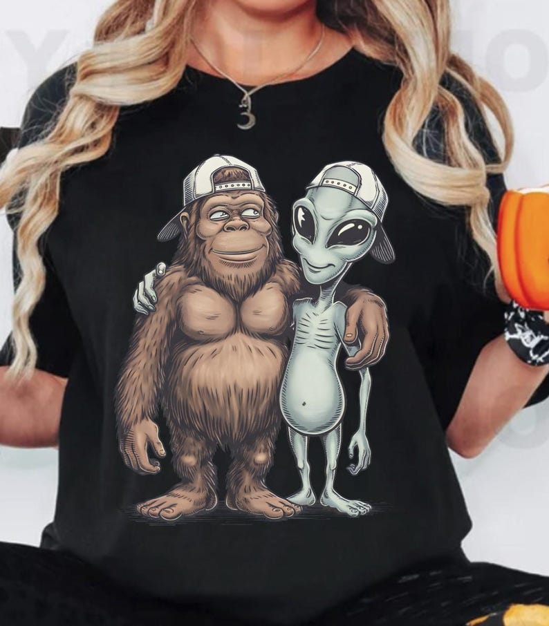 May include: Black t-shirt featuring a cartoon illustration of a Bigfoot character and an alien, both wearing white baseball caps. The Bigfoot is brown and furry, while the alien is gray with large eyes. They are embracing each other.
