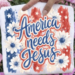 May include: White t-shirt with the text "America needs Jesus" in blue script. The design includes red and blue brushstrokes, white daisies with blue centers, and white stars, creating a patriotic theme.