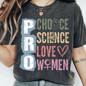 Pode incluir: T-shirt cinzenta escura com as palavras "PRO CHOICE SCIENCE LOVE WOMEN" em várias cores. O design inclui um punho, um símbolo de átomo e um coração. A t-shirt é usada por uma pessoa.