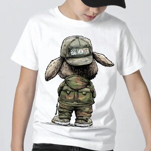 Puede incluir: Camiseta blanca con una ilustraci&oacute;n de dibujos animados de un conejo. El conejo lleva un atuendo de estilo militar y una gorra con la inscripci&oacute;n "EGG HUNTER". La imagen muestra al conejo por detr&aacute;s, con sus orejas y los detalles de su atuendo. La camiseta la lleva una persona.