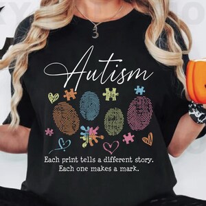 Puede incluir: Camiseta negra con la palabra "Autismo" en escritura blanca. El diseño presenta huellas dactilares de colores, piezas de rompecabezas y corazones. El texto "Cada impresión cuenta una historia diferente. Cada una deja una marca." está impreso debajo del diseño.