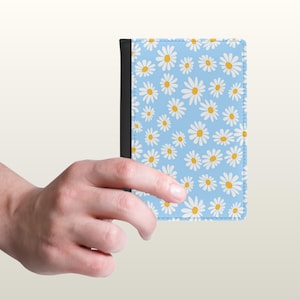 May include: A rectangular passport holder featuring a light blue background with a repeating pattern of white daisies with yellow centers and a solid black spine along the left edge of the cover.