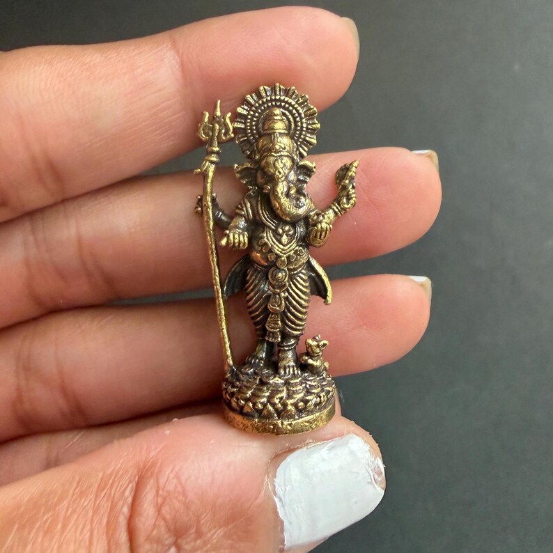 May include: A small, detailed brass statue of the Hindu deity Ganesha. The statue features intricate carvings, a large head, and multiple arms, holding various symbolic objects. Ganesha stands on a detailed base, with a halo behind his head.