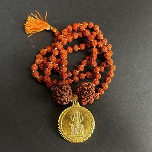 Puede incluir: Un collar mala con cuentas marrones y un colgante dorado. El collar presenta una borla y dos cuentas grandes. El colgante dorado tiene un diseño en relieve. Las cuentas son de color marrón rojizo.