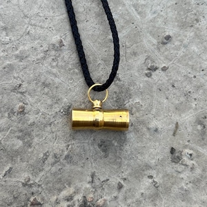 May include: A gold-colored cylindrical pendant hangs from a black braided cord necklace. The pendant has a smooth, polished surface and appears to be made of metal.