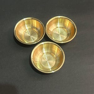 Puede incluir: Tres cuencos pequeños y redondos de latón sobre una superficie oscura. Los cuencos tienen un aspecto dorado brillante con un interior texturizado y un diseño sencillo. Probablemente se utilizan para servir o exhibir. Cada cuenco mide unos 5 cm de diámetro.