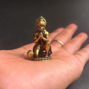 Puede incluir: Pequeña estatua dorada de una figura arrodillada con las manos juntas en oración. La estatua tiene detalles intrincados, incluyendo una corona y una base ornamentada. La figura sostiene una maza. Es un artículo religioso o espiritual.