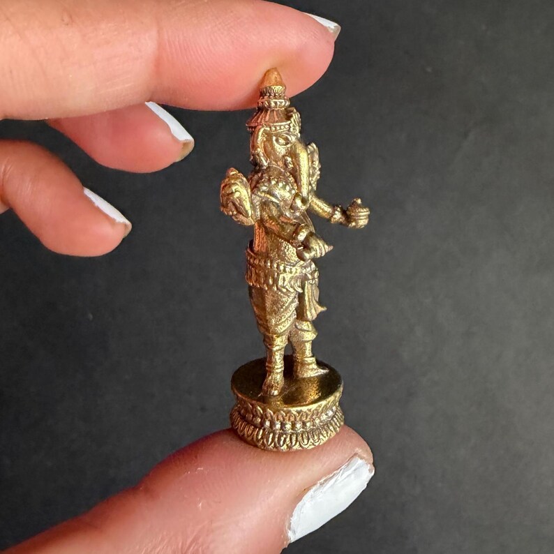 May include: A small, gold-coloured statue of the Hindu deity Ganesha. The statue is intricately detailed, depicting Ganesha with his characteristic elephant head, multiple arms, and traditional attire. The statue is standing on a decorative base.