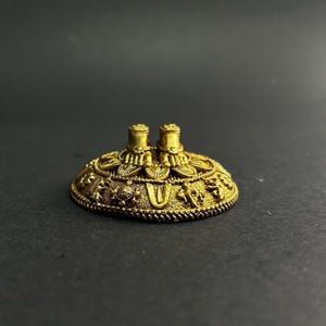 Könnte beinhalten: Ein goldfarbenes, verziertes, kreisförmiges Objekt mit zwei zylindrischen Vorsprüngen. Das Objekt weist detaillierte Reliefs von Figuren und Mustern um seinen Umfang auf. Der Hintergrund ist einfarbig, dunkelgrau.