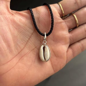May include: A seashell pendant necklace with a black braided cord. The pendant is an oval-shaped, cream-coloured seashell encased in a silver-coloured metal frame. The necklace is displayed on a hand.