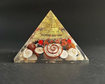 May include: A clear resin pyramid with a gold-colored structure at the top. The pyramid is filled with seashells, red beads, and a copper wire spiral. The base of the pyramid is filled with white seashells.