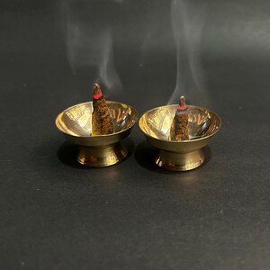 Op de afbeelding: Twee kleine, goudkleurige messing kommen bevatten elk een brandende wierookkegel. De wierookkegels zijn bruin met rode uiteinden en slierten witte rook stijgen op tegen een donkere achtergrond. De kommen hebben een decoratief bladerpatroon.