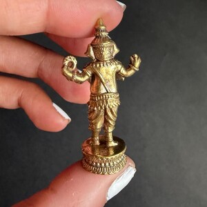 May include: A small, gold-coloured figurine of a deity, possibly a Thai or Southeast Asian figure. The statue is standing on a decorative base and has intricate details in its clothing and headdress. The figurine is held between fingers, showing its small size.