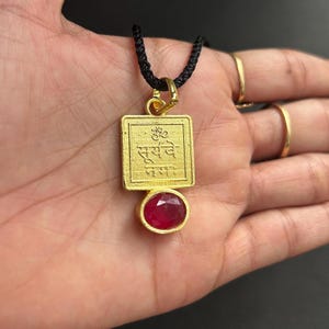 May include: Gold-colored pendant necklace with a rectangular top and a round, red gemstone at the bottom. The pendant has engraved text and is suspended from a black cord.