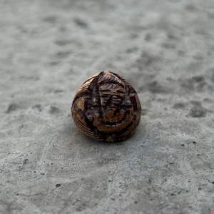 May include: A small, round, brown and red clay bead with intricate carvings. The bead has a textured surface and a geometric design.