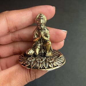 May include: A small, detailed brass figurine of a deity in a kneeling position with hands clasped in prayer. The figure is on a round base with floral and leaf patterns. The figurine has a gold and bronze color.