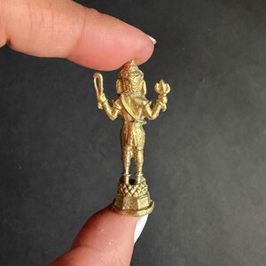 May include: A small, gold-coloured statue of a deity, held between fingers. The statue is detailed, with arms raised, holding objects. It stands on a decorative base. The figure is facing away from the viewer, with intricate details visible.