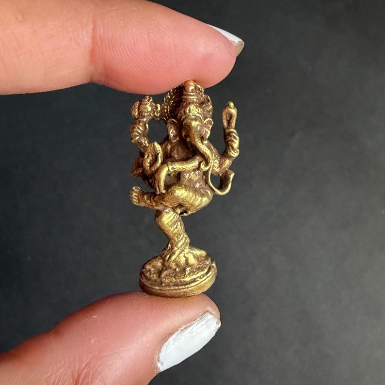 May include: A small, gold-coloured statue of the Hindu deity Ganesha. The figurine is intricately detailed, depicting Ganesha in a dancing pose with multiple arms and a large belly. The statue is held between two fingers against a dark background.