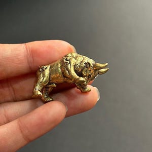 May include: A small, detailed brass figurine of a bull in a charging pose. The bull has prominent horns and a textured body, with a golden-yellow color. The figurine is held in a person's hand against a gray background.