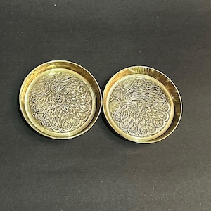 May include: Two brass coasters featuring a detailed peacock design. The coasters are circular with a raised edge. The peacock design is in the centre of each coaster, with intricate feather details.