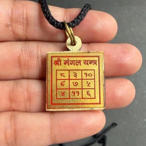 Peut inclure: Un pendentif carré de couleur or avec un motif et un texte rouges et dorés. Le pendentif est attaché à un cordon noir. Le texte sur le pendentif est dans une langue étrangère. Le pendentif est tenu par une personne.