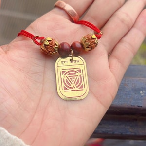 Op de afbeelding: Een goudkleurige hanger met de tekst "दुर्गा कवच" en een geometrisch ontwerp, geregen aan een rood koord met bruine kralen en decoratieve elementen. De hanger is rechthoekig met afgeronde hoeken.