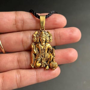 May include: Gold-coloured pendant necklace featuring a detailed depiction of a deity with multiple faces. The pendant is suspended from a black cord. The deity is holding a mace. The pendant is held in a person's hand.