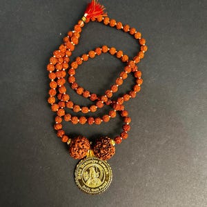 May include: A necklace with reddish-brown beads and a gold-coloured pendant. The beads are strung together, forming multiple strands. The pendant has an image and text. A red tassel hangs from the top of the necklace.