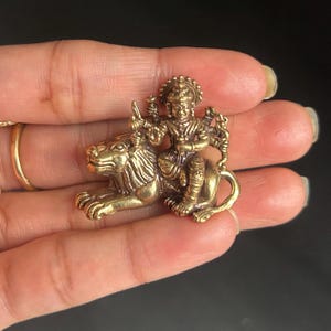 May include: A small, detailed brass figurine depicting a deity riding a lion. The figure is adorned with intricate details, including a crown and multiple arms holding various objects. The lion is also intricately detailed, with a flowing mane and a powerful stance.