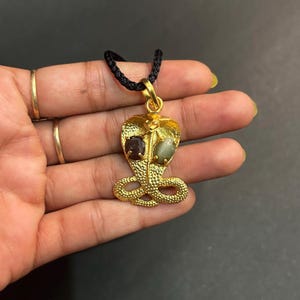 Könnte beinhalten: Goldfarbener Cobra-Anhänger an einer schwarzen Kordel. Der Anhänger zeigt ein Cobra-Kopf-Design mit zwei eingesetzten Edelsteinen, einem dunkelroten und einem hellgrünen. Der Körper der Kobra ist mit strukturierten Schuppen versehen.