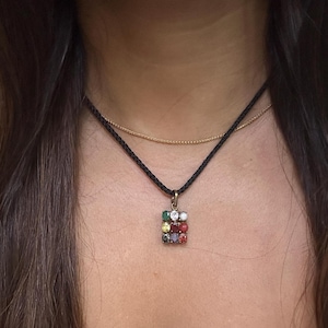 May include: A gold-toned pendant necklace with a square, multi-colored gemstone design. The pendant is suspended from a black cord necklace, with a delicate gold chain layered above. The gemstones are arranged in a grid pattern.