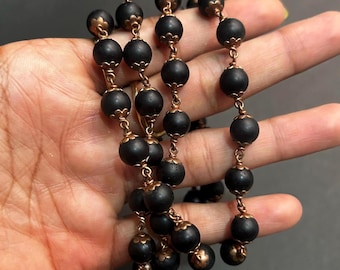 May include: A vintage necklace featuring matte black beads strung together with ornate copper-colored metal links. The beads are round and evenly spaced, creating a classic and elegant design. The necklace is held in a hand, showcasing its length and detail.
