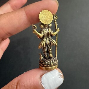 May include: A small, detailed brass figurine of a deity, with a sun-like halo and holding a trident and a small object. The figure stands on a detailed base, with a small animal at its feet. The figurine has an aged, antique appearance.