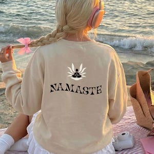 Puede incluir: Una sudadera beige con la palabra "NAMASTE" impresa en negro, combinada con una falda blanca. La imagen muestra a una persona con auriculares rosas, sentada en una manta a rayas cerca del mar. Un lazo rosa adorna el cabello.