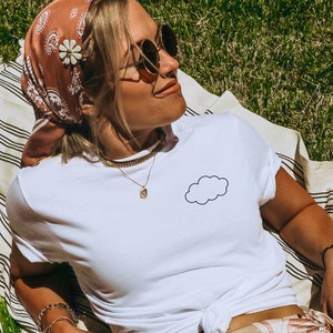 Könnte beinhalten: Eine Frau trägt ein weißes T-Shirt mit einem kleinen Wolken-Motiv auf der Vorderseite. Sie trägt einen braunen und weißen gemusterten Rock und ein braunes Bandana mit einer weißen Blume.