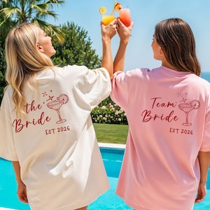 Può includere: Due donne indossano magliette oversize con grafiche di cocktail e testo. Una maglietta è color crema con "the Bride" e l'altra rosa con "Team Bride". Entrambe le magliette hanno "EST 2026" stampato sotto. Tengono cocktail a bordo piscina.