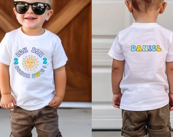 May include: A white t-shirt with the text "ICH BIN 2 SCHON ZWEI" and a sun graphic. The back of the shirt has the name "DANIEL" in colorful letters. The shirt is worn by a young child with sunglasses and brown shorts.