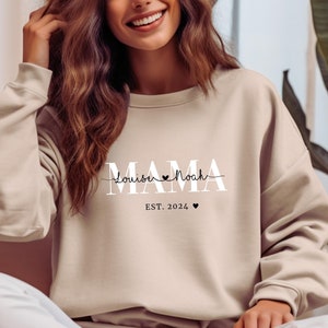 Könnte beinhalten: Ein beiger Pullover mit dem Text "MAMA" in weißen Buchstaben, gefolgt von "louisa Noah" in kursivschrift mit einem Herzsymbol. Der Text "EST. 2024" befindet sich unter dem kursivschriftlichen Text mit einem kleinen Herzsymbol.