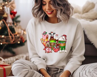Peut inclure: Sweat-shirt blanc avec un motif de Noël représentant des animaux de ferme portant des bonnets de Père Noël et tenant des tasses. Les animaux comprennent un cochon, une vache, un lama, un mouton et une oie.