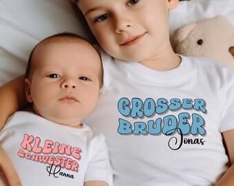May include: Two children wearing white shirts. The older child's shirt says "GROSSER BRUDER Jonas" in blue bubble letters. The younger child's shirt says "KLEINE SCHWESTER Hanna" in pink bubble letters. A teddy bear is in the background.