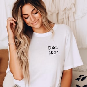 Może przedstawiać: Biała koszulka z napisem "DOG MOM" w kolorze czarnym, z motywem łapy. Koszulka ma swobodny styl, z kr&oacute;tkimi rękawami. Zdjęcie przedstawia koszulkę na osobie.
