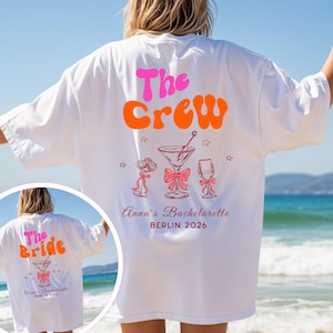 Op de afbeelding: Wit oversized T-shirt met de tekst "The Crew" in roze en oranje, cocktail illustraties en de tekst "Anna's Bachelorette BERLIN 2026". Het shirt wordt op het strand gedragen.