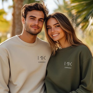Op de afbeelding: Twee mensen dragen bijpassende sweatshirts met de tekst "11K 22092022" op de voorkant. De trui aan de linkerkant is beige en de trui aan de rechterkant is groen.