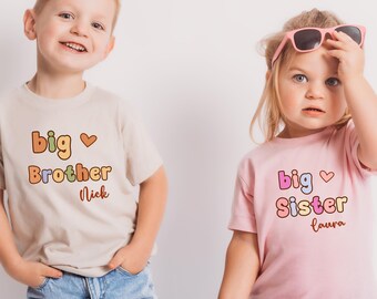 May include: Two children wearing matching t-shirts. One shirt is tan with the words "Big Brother Nick" in orange and brown. The other shirt is pink with the words "Big Sister Laura" in pink, purple, and yellow. Both shirts have a heart symbol.