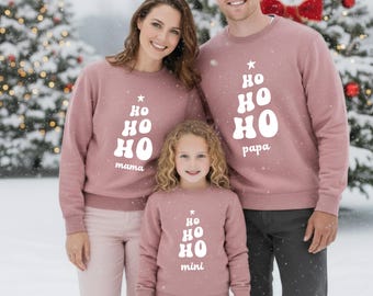 May include: A family wearing matching mauve sweatshirts with a Christmas tree design and the words "Ho Ho Ho." The sweatshirts have the words "mama," "papa," and "mini" printed on them. The family is standing in front of a snowy Christmas tree background.