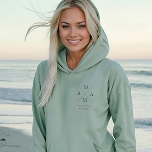 Peut inclure: Un sweat-shirt à capuche vert clair avec un motif graphique noir sur le devant. Le graphique présente les lettres "M A M A" disposées en forme de croix avec le texte "Leonardo est 2024" en dessous.