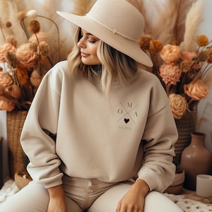 Op de afbeelding: Een beige sweatshirt met een zwart grafisch ontwerp met de letters "OMA" in een kruisvorm met een hart in het midden. De tekst "Est. 2023" staat onder de grafiek.