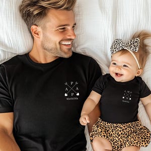 Könnte beinhalten: Schwarze T-Shirts mit passenden Designs. Ein Shirt hat "P A P A" mit Pfeilen und Herzen und den Namen "Mathilda" darunter. Das andere Shirt hat "M A M I" mit Pfeilen und Herzen und den Namen "Mathilda" darunter.