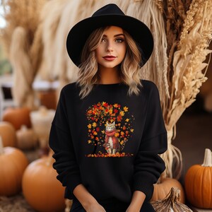 Könnte beinhalten: Schwarzes Sweatshirt mit einem Aufdruck eines Katers mit Schal, der in einem Baum mit Herbstlaub steht.